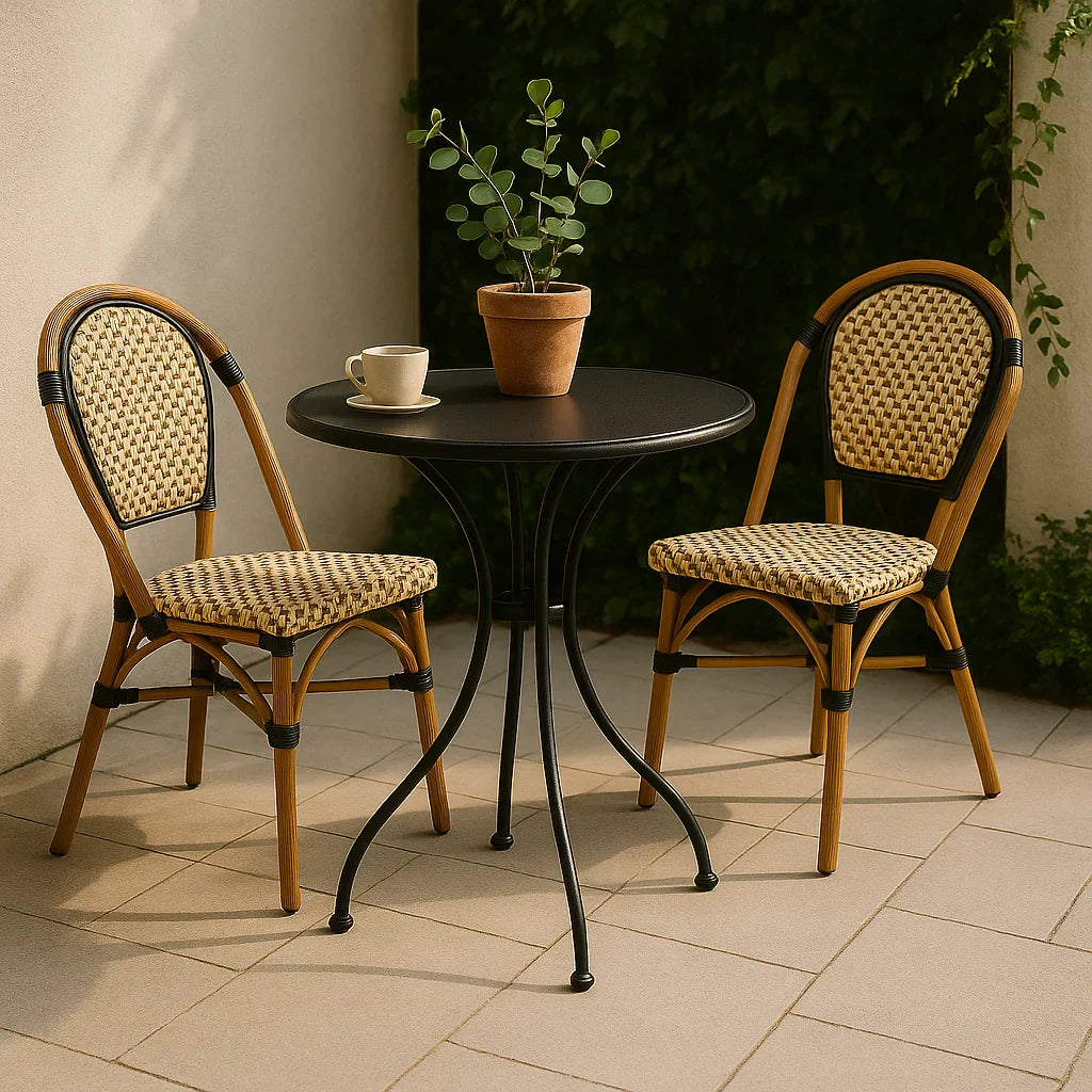 Franse bistroset met zwart metalen rond tafeltje en twee gevlochten rattan stoelen op een zonnig terras, naast een terracotta pot met plant.