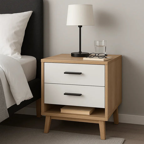 Modern nachtkastje van hout met twee lades en een open vak, geplaatst naast een bed met grijze stoffering en witte dekens, voorzien van een zwarte tafellamp en een glas water erbovenop.