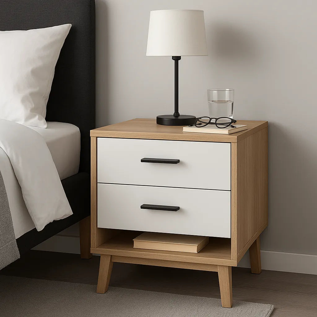 Modern nachtkastje van hout met twee lades en een open vak, geplaatst naast een bed met grijze stoffering en witte dekens, voorzien van een zwarte tafellamp en een glas water erbovenop.