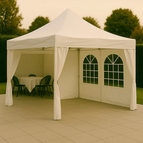 Witte partytent met puntdak en boogvormige ramen, geplaatst op een betegeld terras in een groene tuin. 