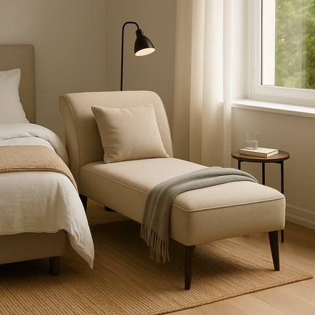 Beige chaise longue naast bed in lichte slaapkamer met zacht daglicht, plaid en bijzettafeltje met glas water.